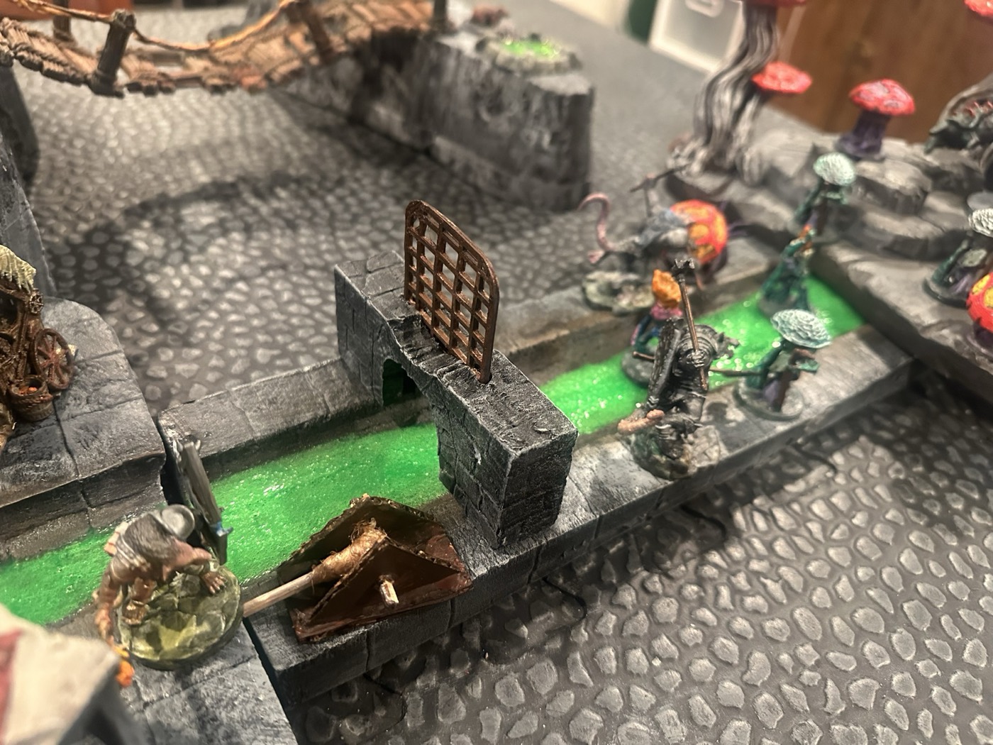 Miniatures of an adventuring party wading along a green sewer channel toward a grated arch, with a rope bridge and giant mushrooms behind them.